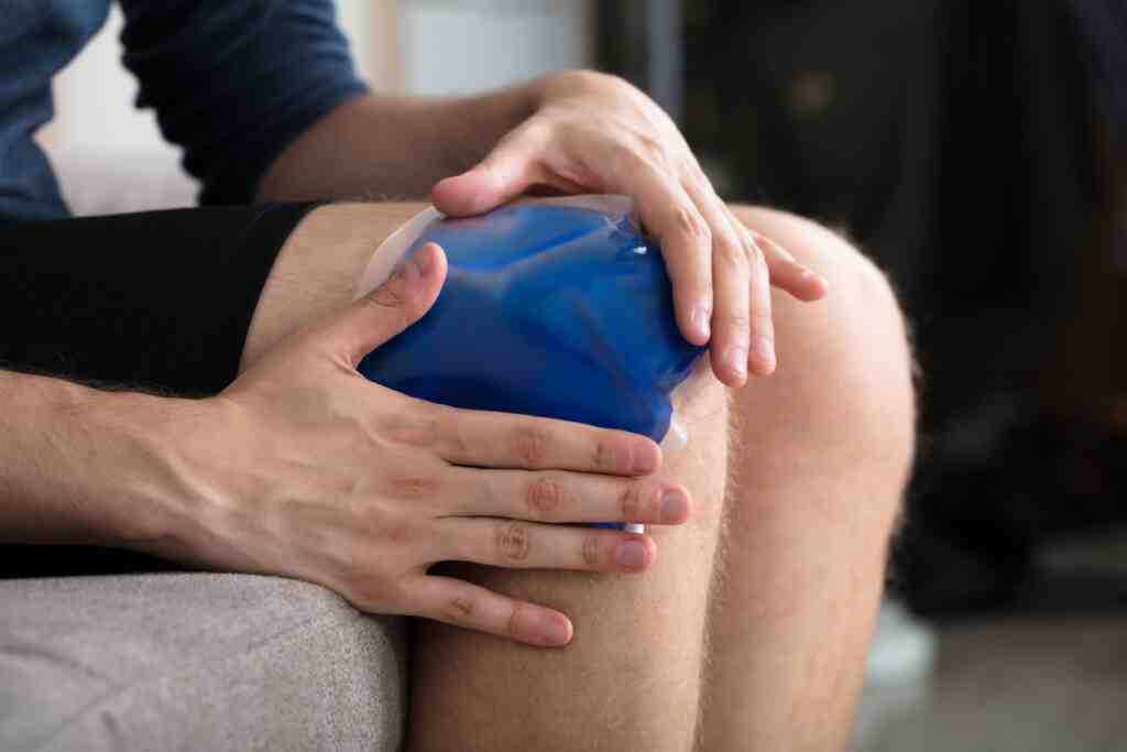 A person sits on a couch, applying a blue ice pack to their knee
