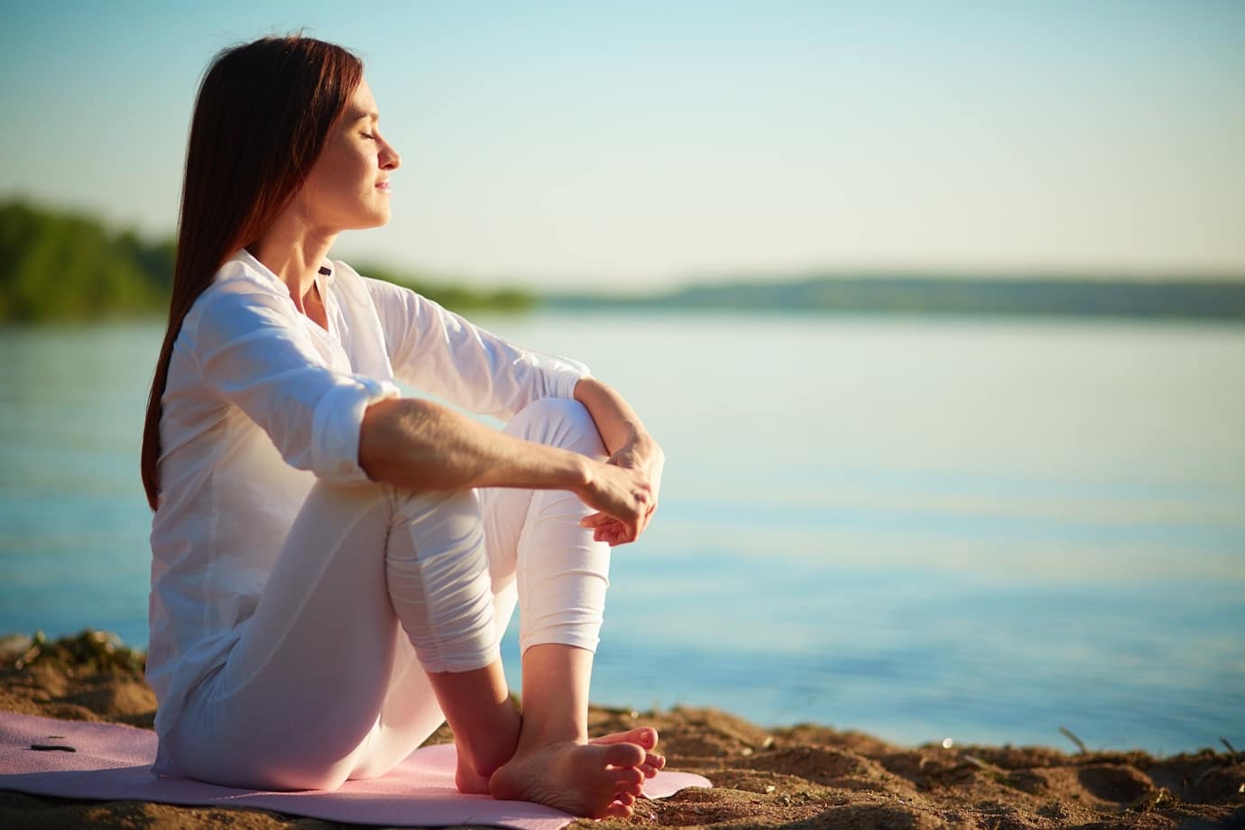 woman in white clothing sits meditatively by a lake
