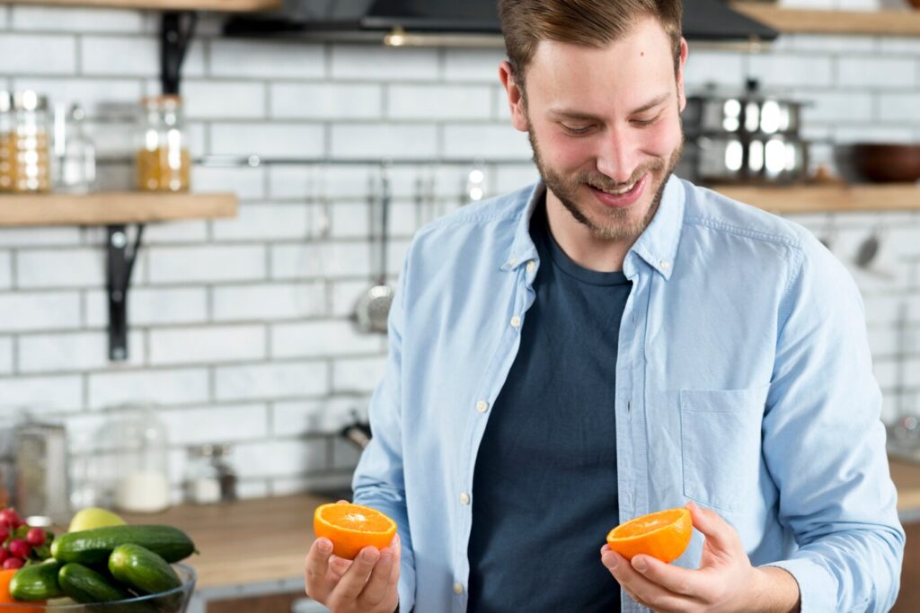 man in a kitchen holds two halves of an orange, surrounded by fresh vegetables