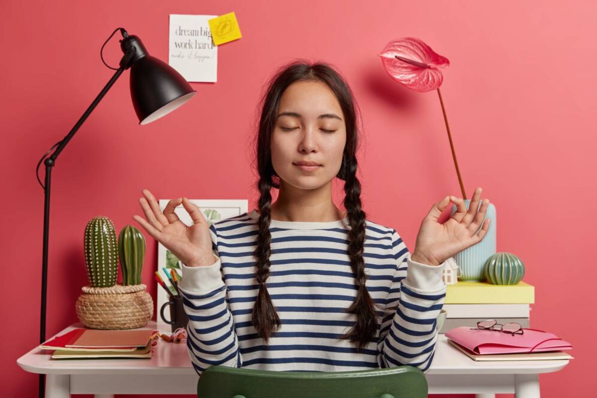 Young woman meditating at a desk with a striped shirt, surrounded by desk items and a pink background.
