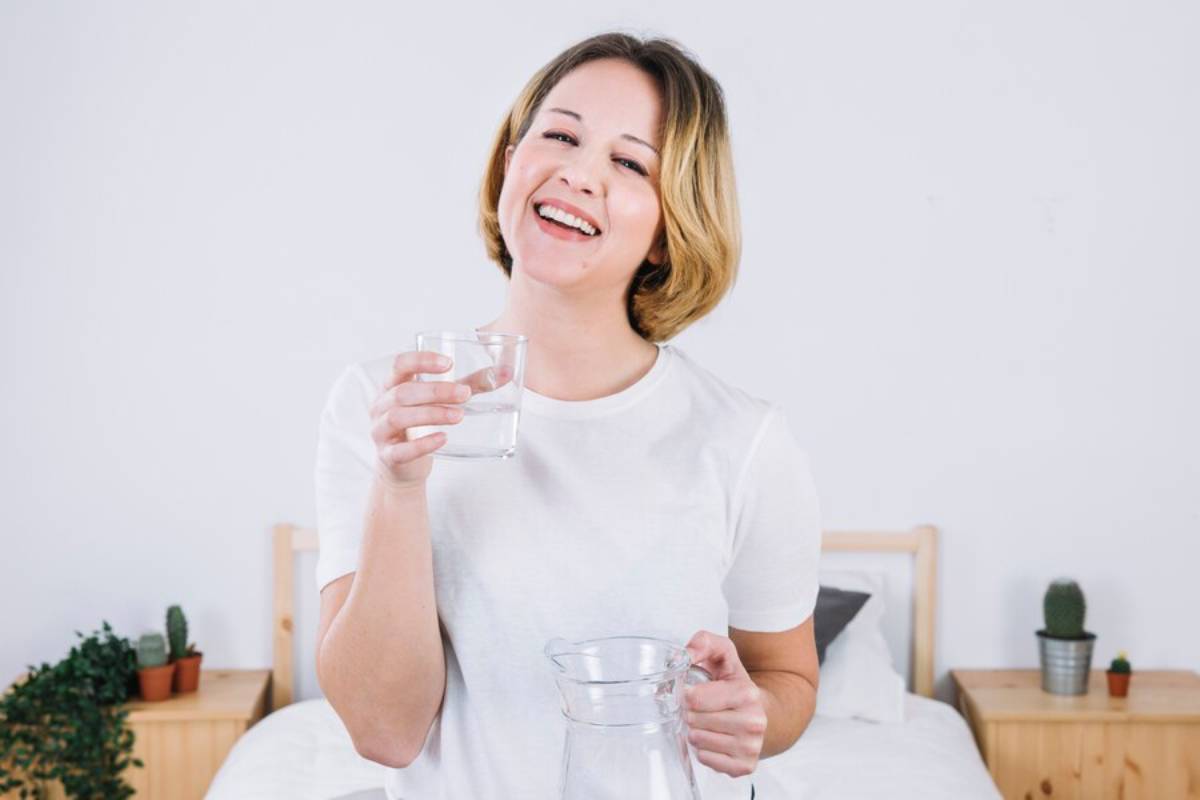 Smiling woman holding a glass of water in a bedroom with plants on the background.