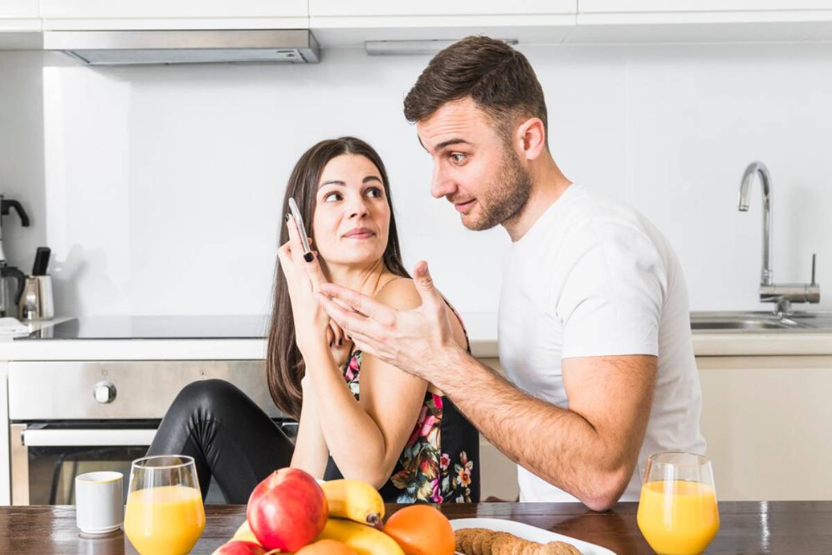 Man and woman talking in kitchen; woman holds smartphone; breakfast table with fruit and orange juice visible.