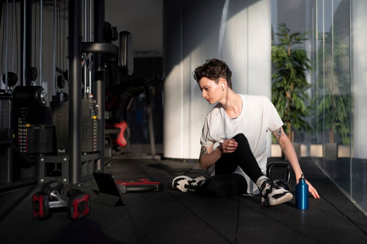 Young man with earphones sitting on gym floor, resting and looking at a tablet beside workout equipment.