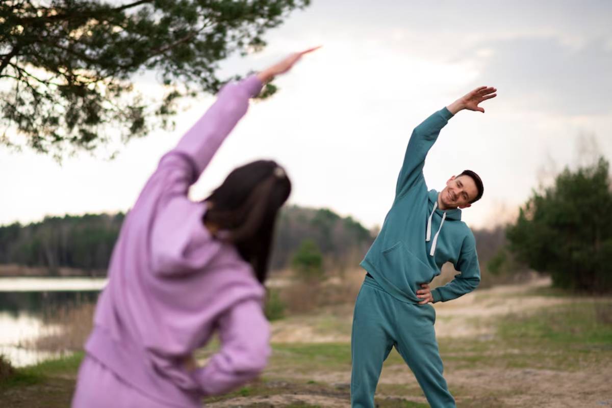 Two people stretching outdoors near a lake, one in purple and the other in teal, engaging in a morning exercise routine.
