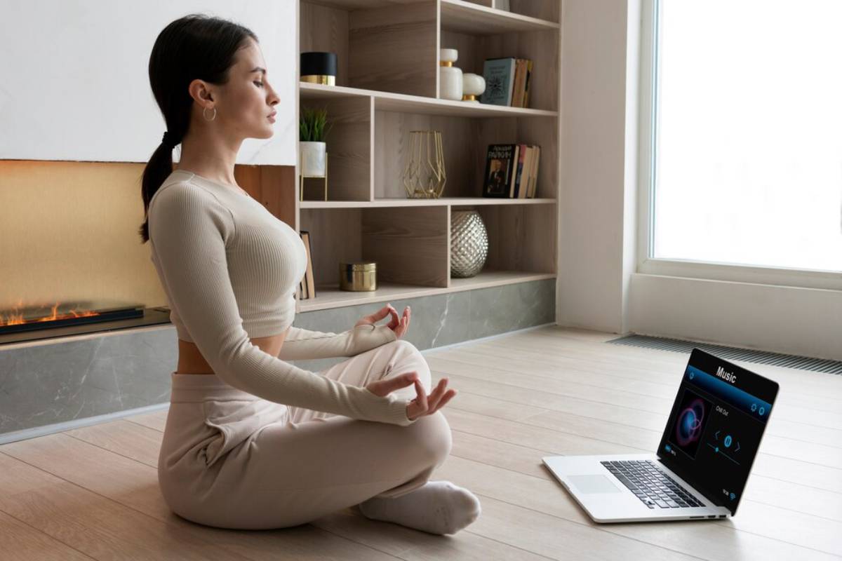 Woman in beige activewear meditating on a yoga mat, using a laptop with a music app open, in a modern living room.