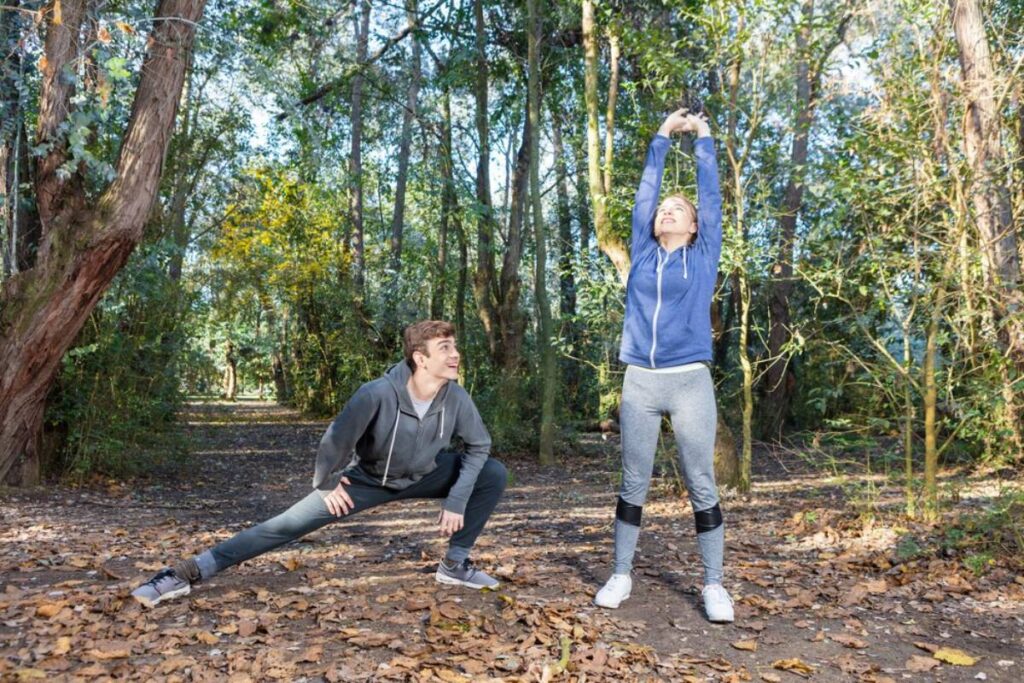 Two young adults in sportswear stretching in a sunlit forest, surrounded by tall trees and fallen leaves.