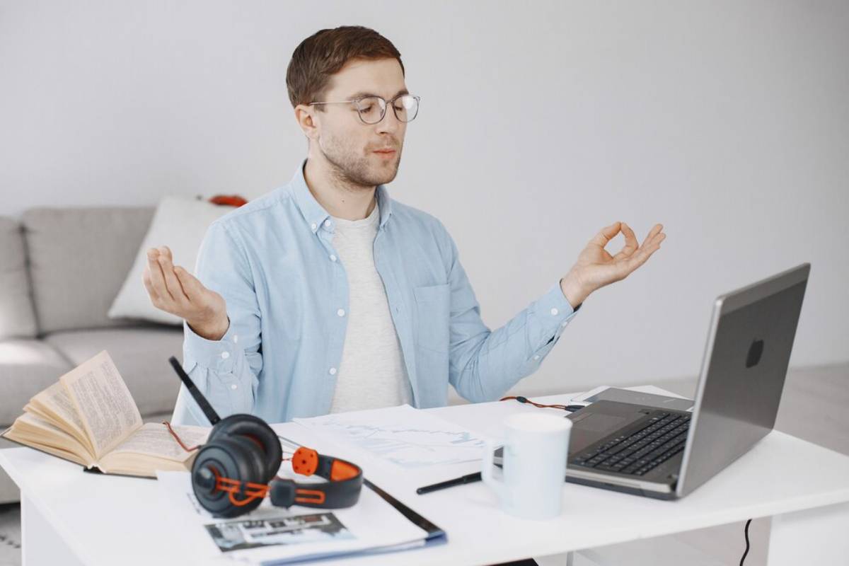 Man meditating at desk with laptop, books, and headphones, displaying a peaceful expression.