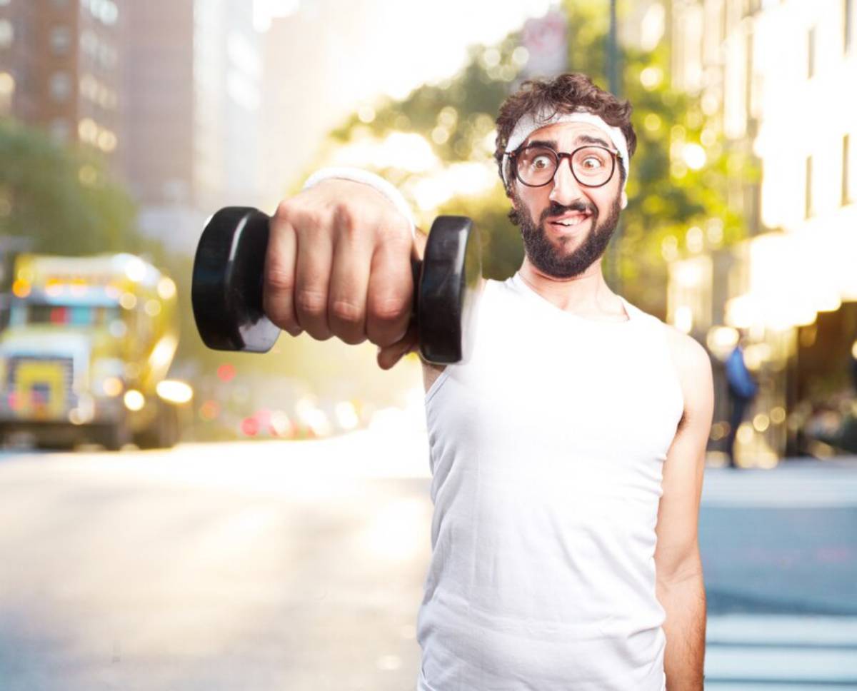 Man in white tank top and glasses lifting a dumbbell, smiling, with a city street background.