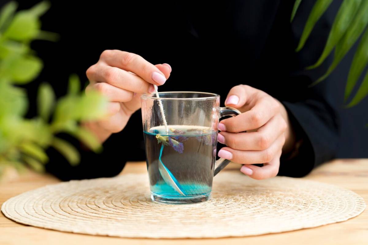 Person steeping a blue flowering tea in a clear glass mug on a wooden table, surrounded by green plants.