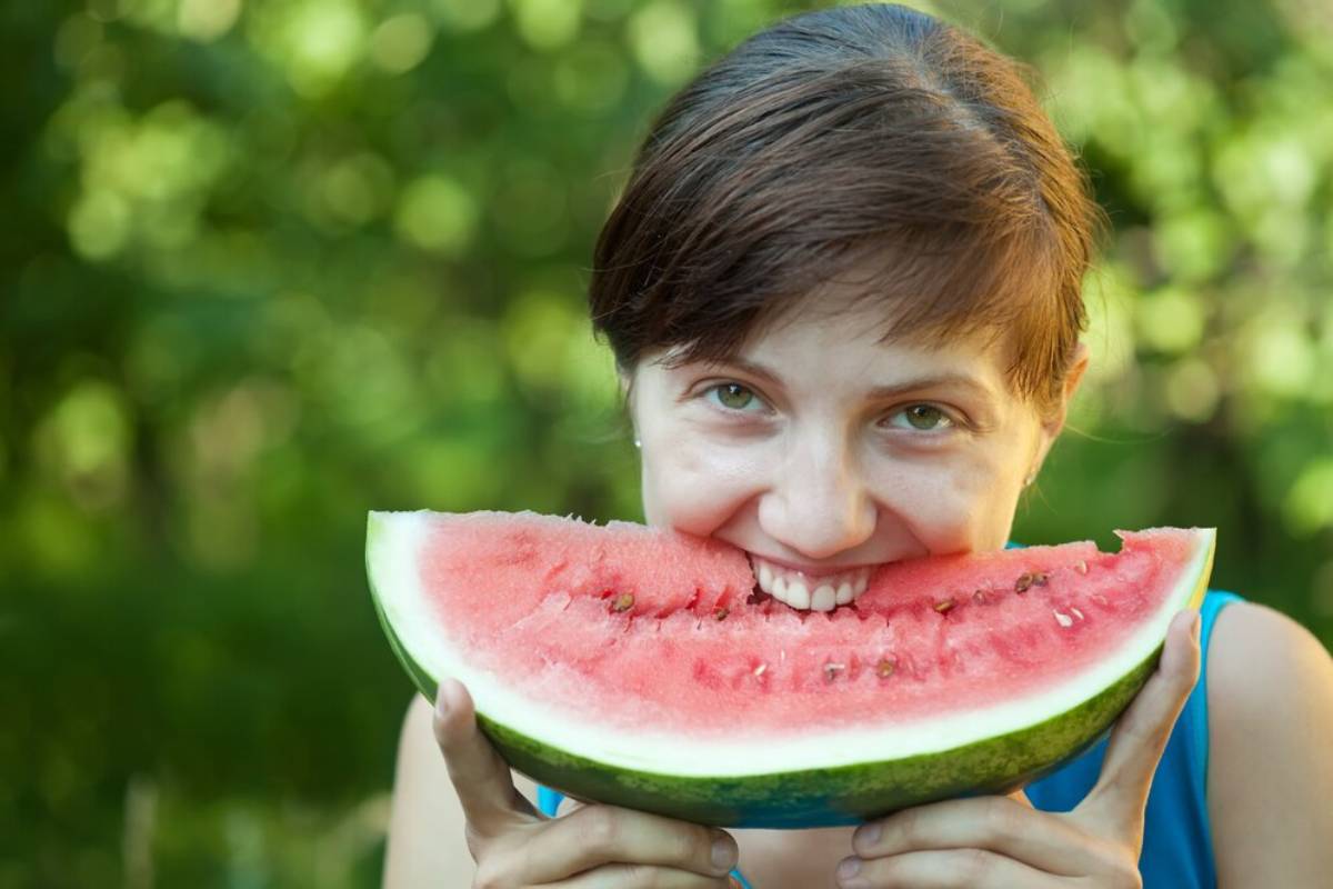 Woman playfully biting into a large slice of watermelon, outdoors with a blurred green background.